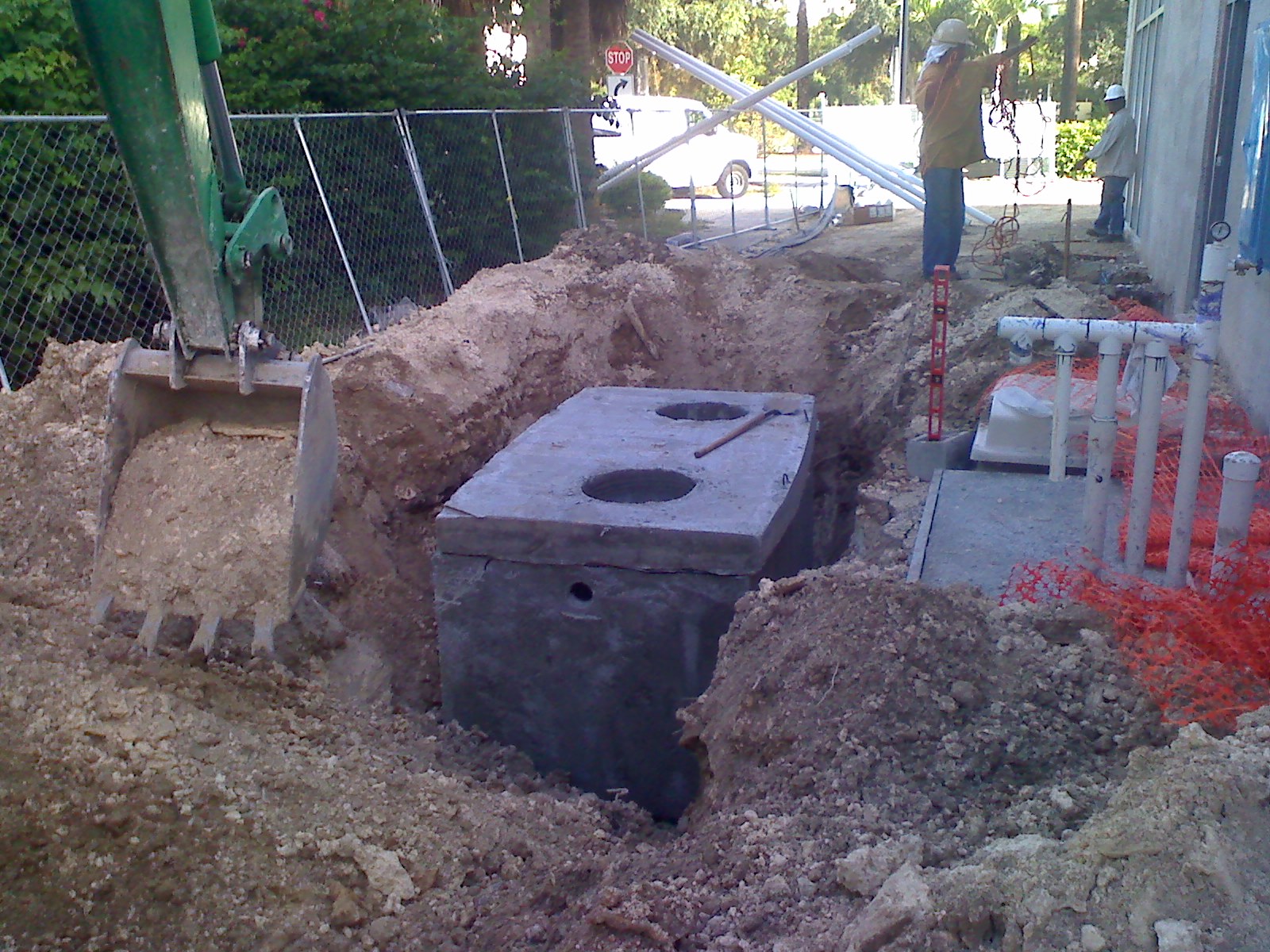 Excavator lowering a concrete septic tank into an open pit at an active install site