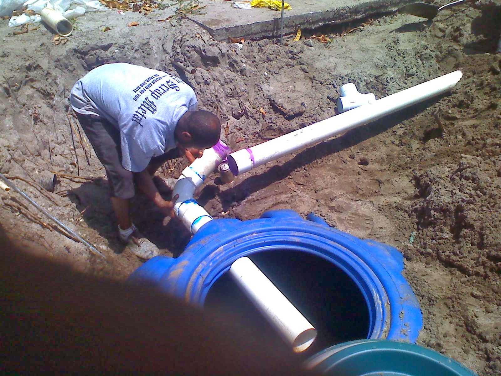 Technician connecting PVC line to a blue plastic septic tank in a trench